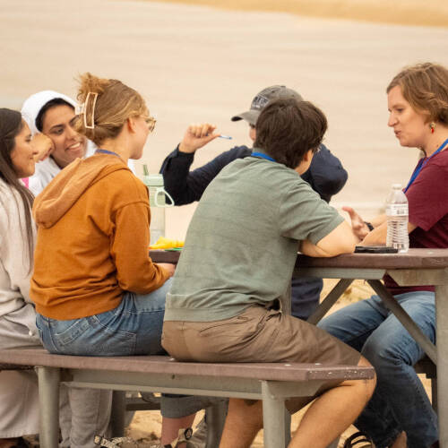 A group of people at a picnic table on the beach chatting and smiling.